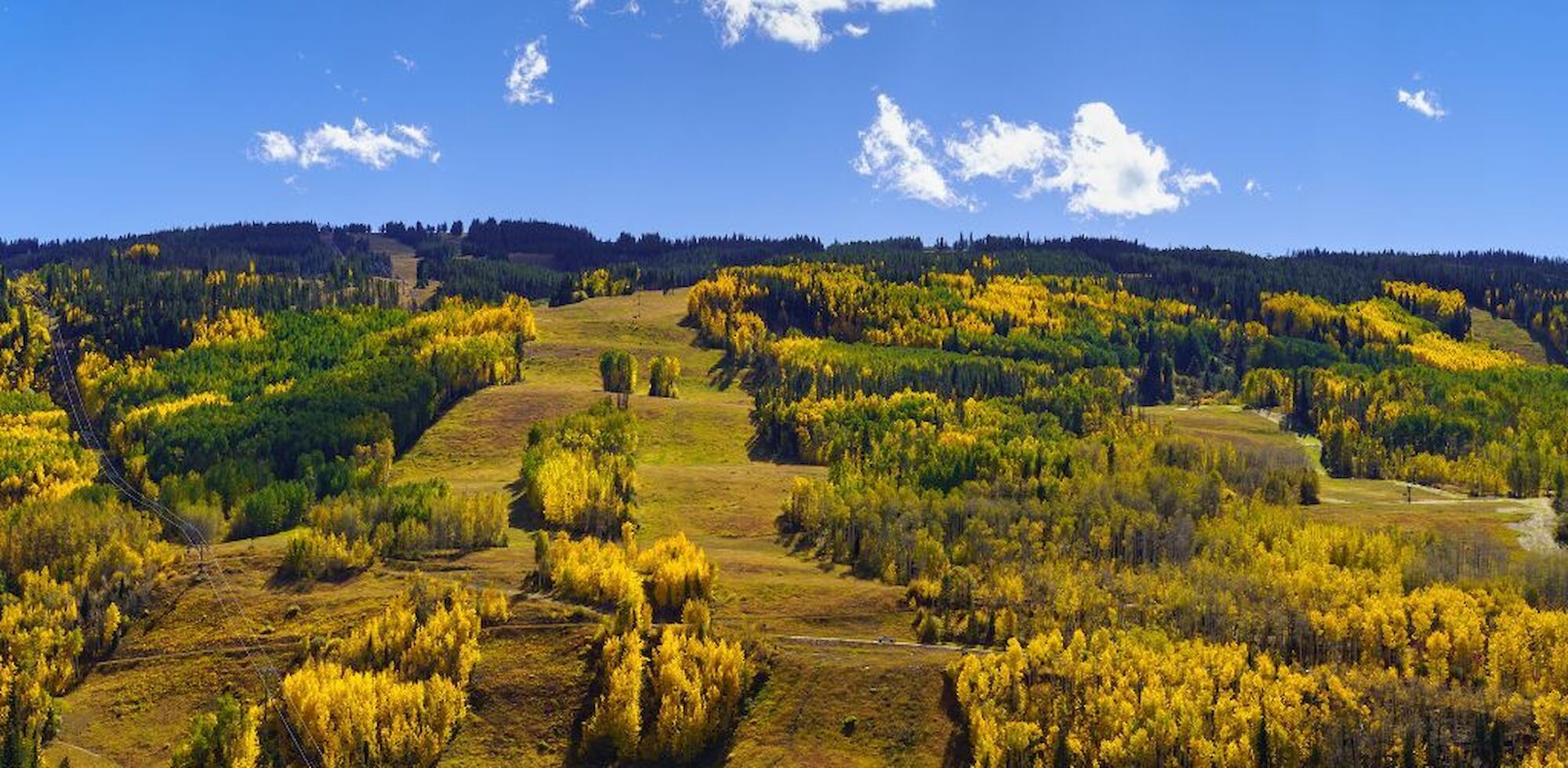 A scenic view of a mountainous area with lush autumn foliage and a small town nestled at the foothills under a bright blue sky with scattered clouds.