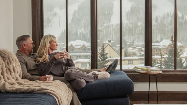 A couple enjoys a cozy moment together on a couch, with a snowy mountain view outside their large windows.
