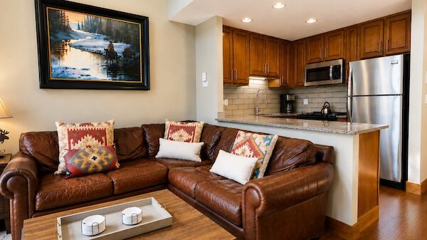 Cozy contemporary living area with a brown leather sectional, colorful throw pillows, framed landscape art, and an open kitchen with stainless steel appliances.