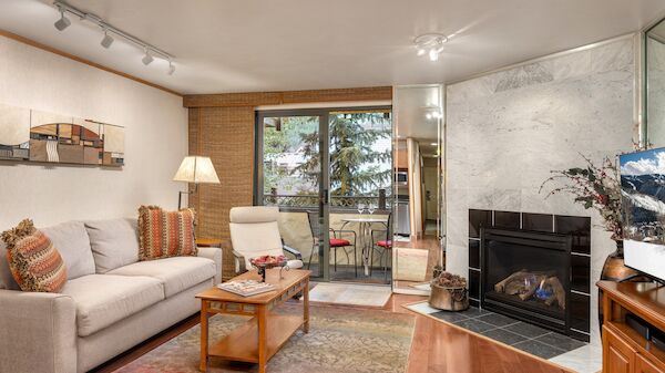 Cozy living room with a beige sofa, wooden coffee table, fireplace, and a view to a small balcony with plants; warm lighting and decorative rug trail.