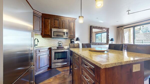 The image shows a modern kitchen with wooden cabinets, stainless appliances, a granite island, and a view through a large window.