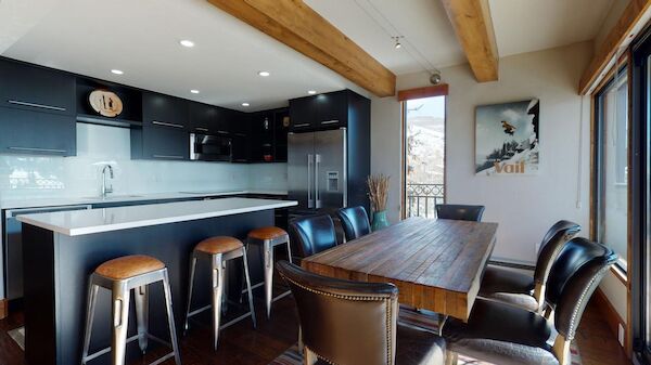 A modern kitchen with dark cabinets, an island with high chairs, a wooden dining table, and large windows providing natural light.