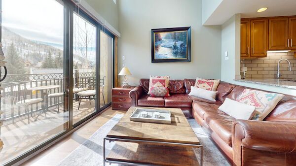 The image shows a cozy living room with a leather sofa, a wooden coffee table, and large windows revealing a beautiful outdoor view.
