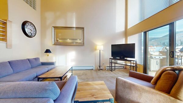 A modern living room with a blue sectional, tan chair, coffee tables, a wall clock, floor lamp, flat-screen TV, and large windows with a balcony view.