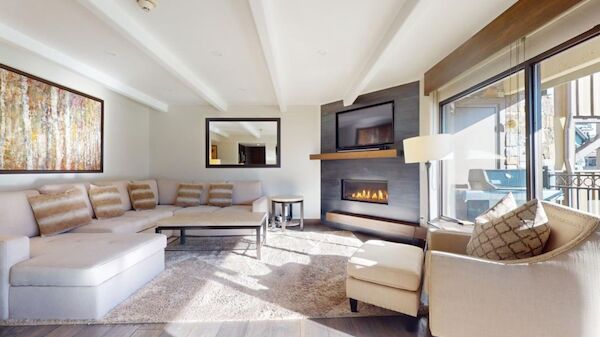 A bright, modern living room with a light beige sectional, matching chairs, a fireplace, a wall-mounted TV, and a large window/slider letting in natural light.