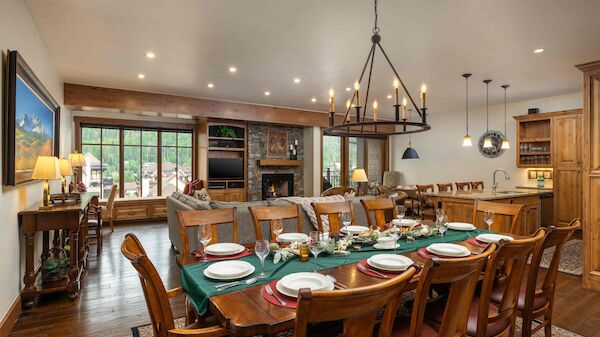The image shows a cozy dining area and living space with wooden furniture, a chandelier, and scenic views outside.