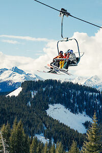 Three skiers ride a chairlift ascending a snowy mountain with pine trees and stunning peaks in the background.