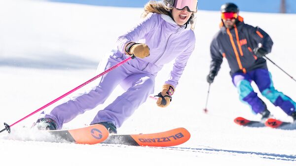 Two skiers descend a snowy slope, one in a lavender outfit leading as the other follows, both carving turns under a clear blue sky.