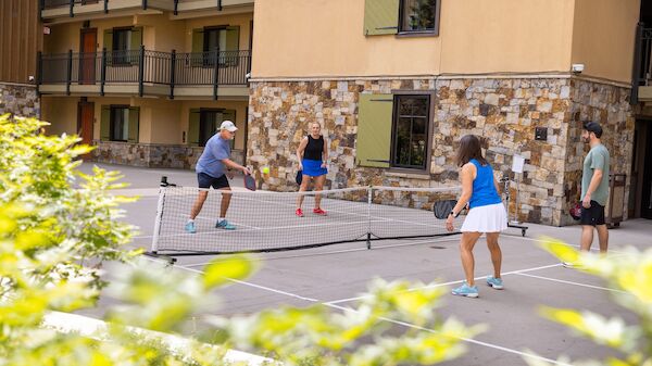 People play pickleball on a sunny courtyard court with a net, surrounded by a stone and stucco building, and green plants in the foreground.