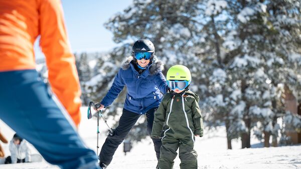 A snowy scene with a snowboarder and a child in protective gear learning to ski, while an instructor approaches from the foreground.