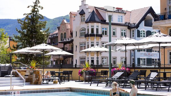 A resort-style pool area with sun loungers and umbrellas, colorful European-style buildings in the background, and people swimming.