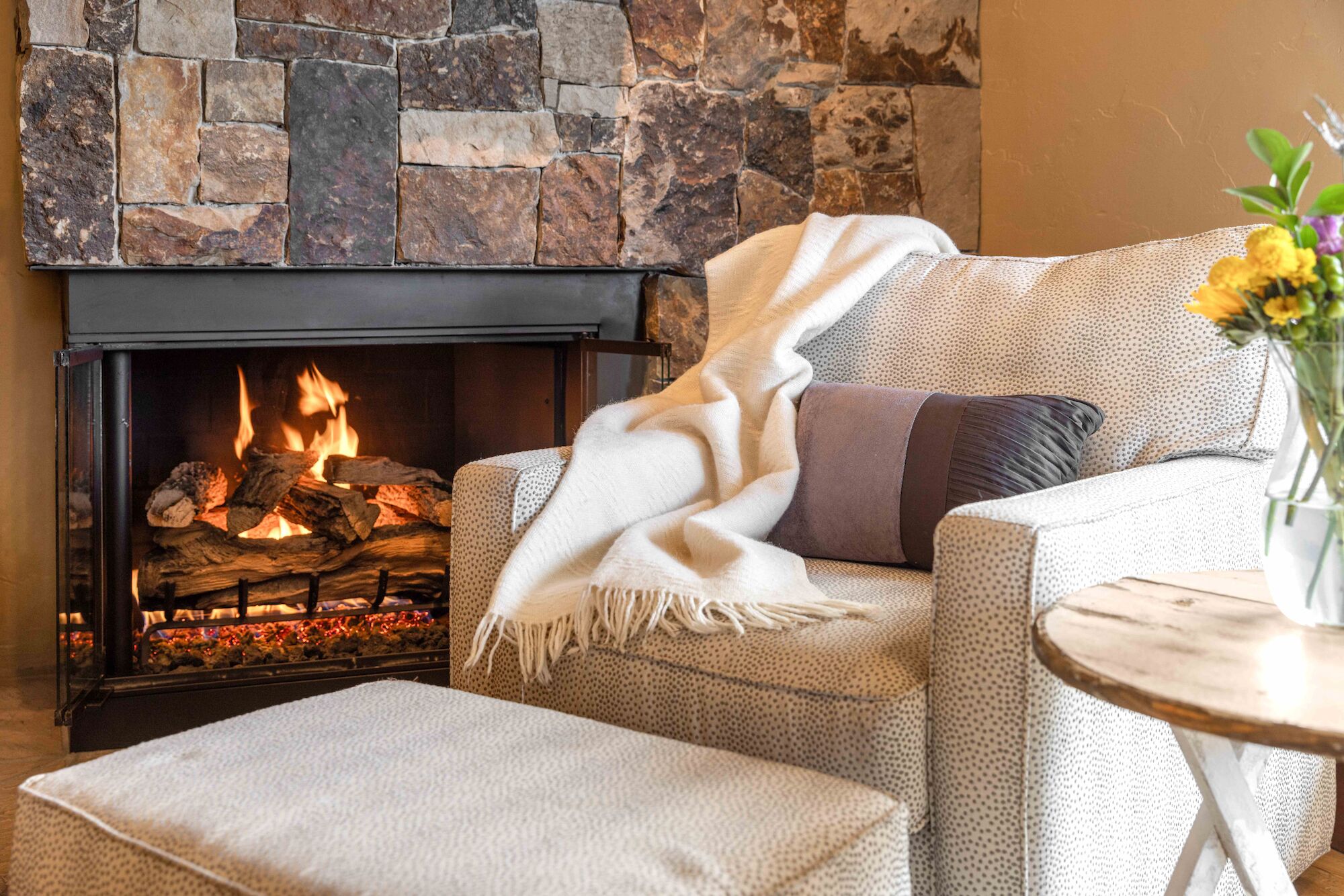 The image shows a cozy living room scene with a fireplace, a comfortable chair draped with a blanket, and a small table with flowers.
