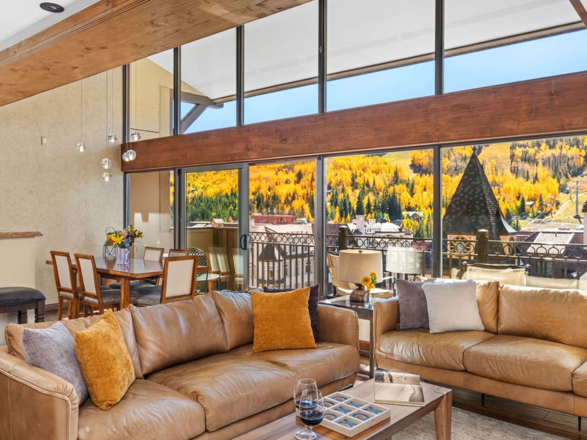 Luxurious open-plan living room with tan leather sofas, mustard cushions, wood beams, and large glass doors overlooking a scenic outdoor patio and autumn trees.