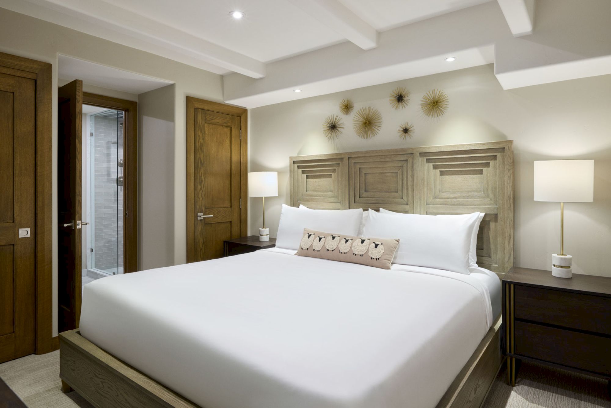 A modern bedroom with a large bed, white linens, decorative pillows, bedside tables with lamps, and wall art. There is an open door to a bathroom.