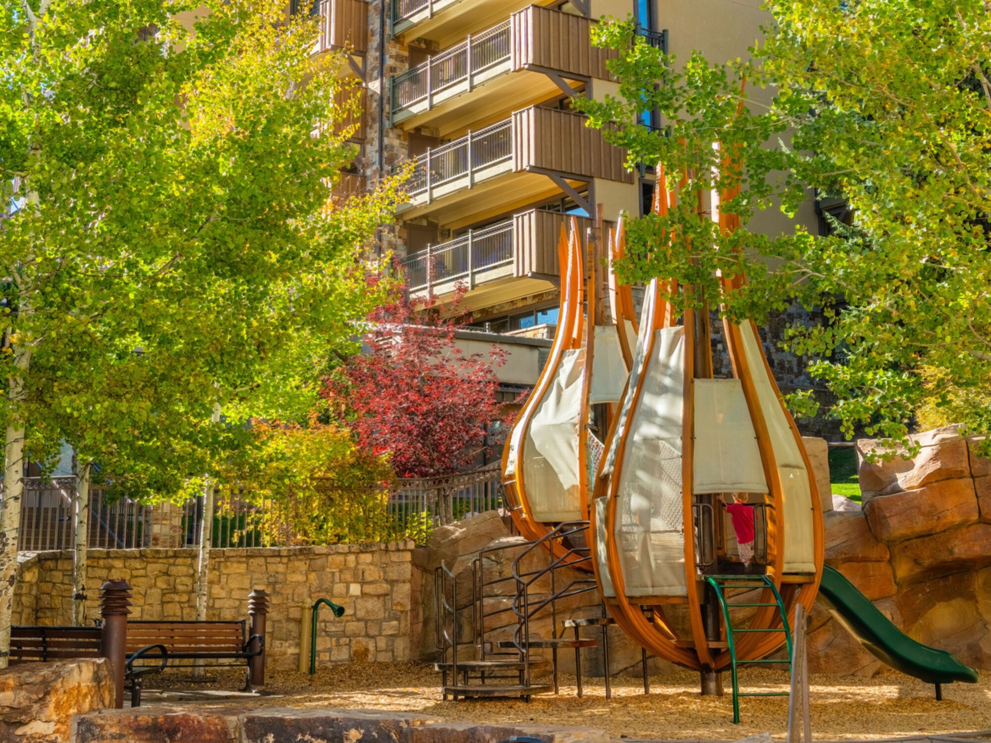 Children&rsquo;s playground with a slide and climbing structure, set against trees and an apartment building in the background.