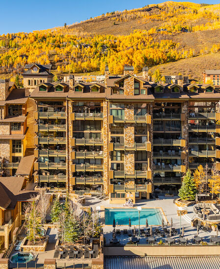 A modern apartment complex with a rooftop pool, surrounded by autumn trees and alpine hills in the background.