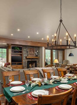 A spacious rustic dining room with a long wooden table set for a meal, warm lighting, a stone fireplace, and a cozy kitchen area in the background.