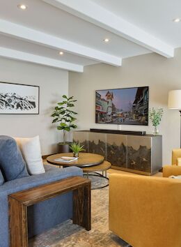 A modern living room with a blue sofa, yellow armchairs, a wooden coffee table, wall-mounted TV, and a few plants in a bright, airy space.