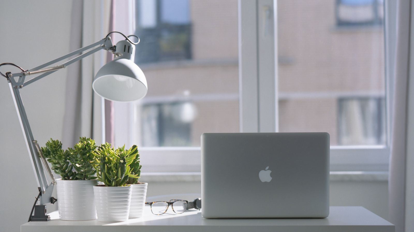 A desk with a laptop, a lamp, two potted plants, and a pair of glasses in front of a window with a building view is depicted.