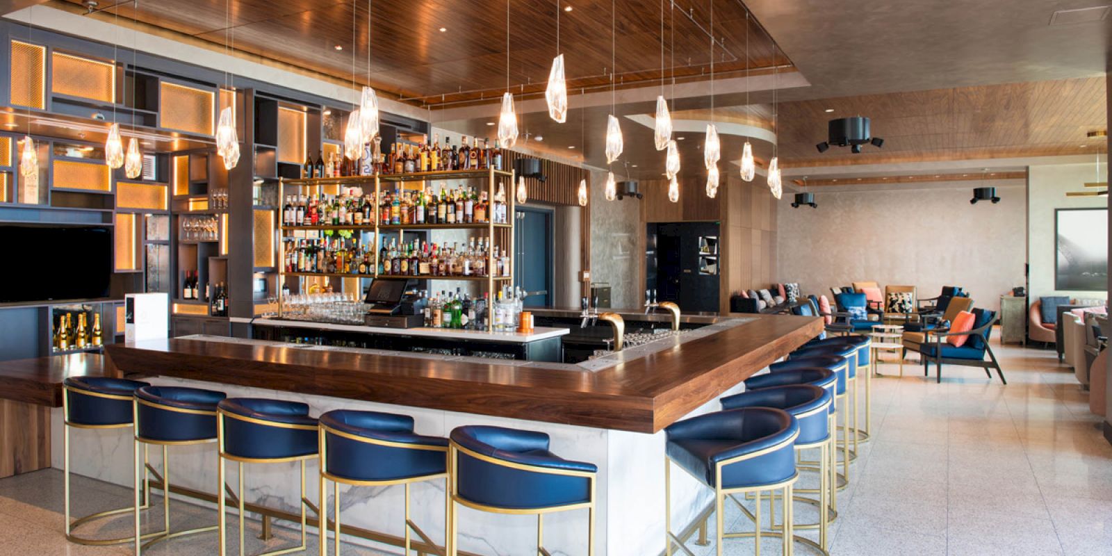 A modern bar with blue and gold seating, a well-stocked bar, and contemporary lighting. Cozy seating is available in the background.