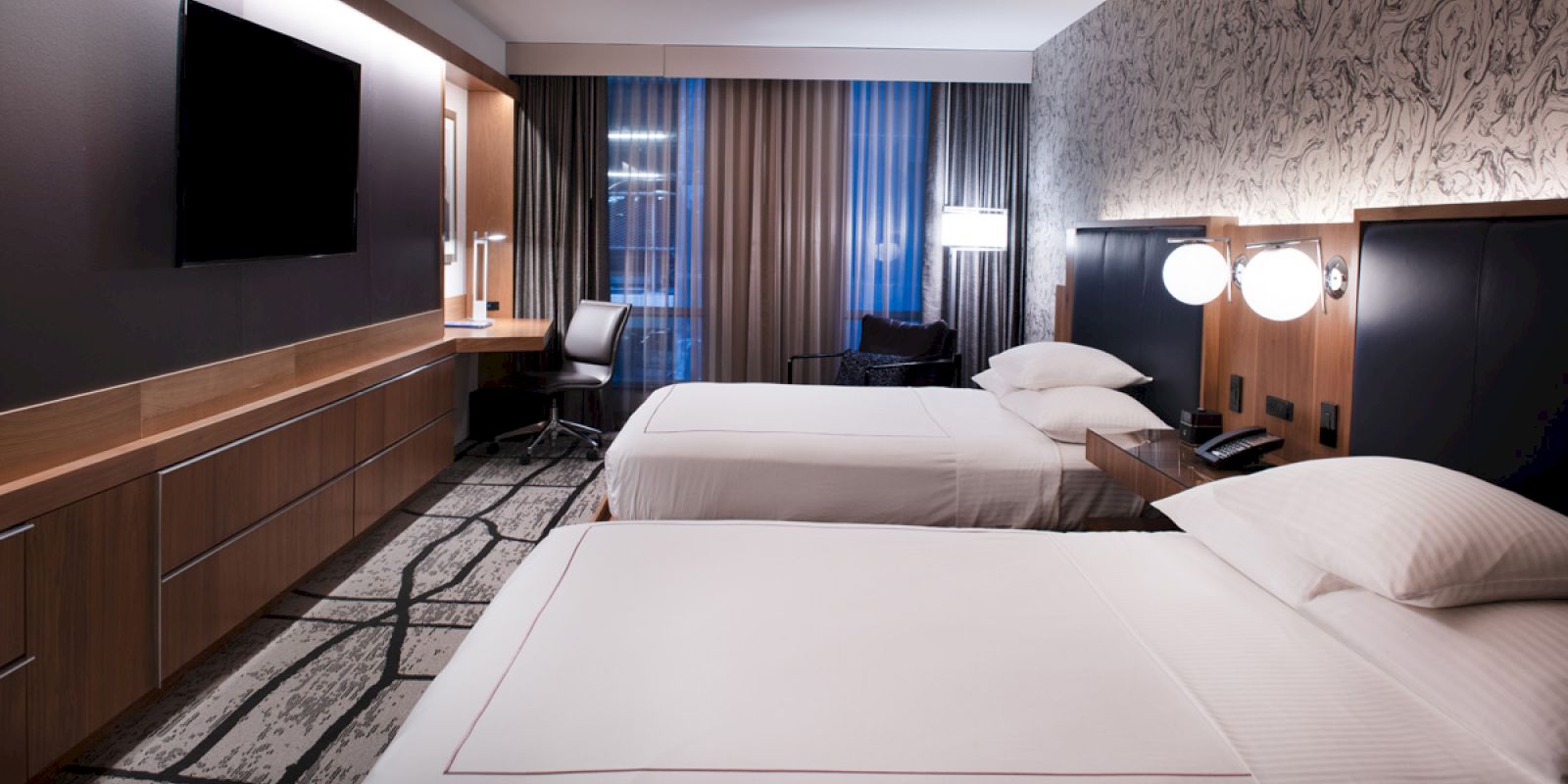 The image shows a modern hotel room with two beds, a wall-mounted TV, a desk with a chair, and contemporary decor. The room is well-lit and stylish.