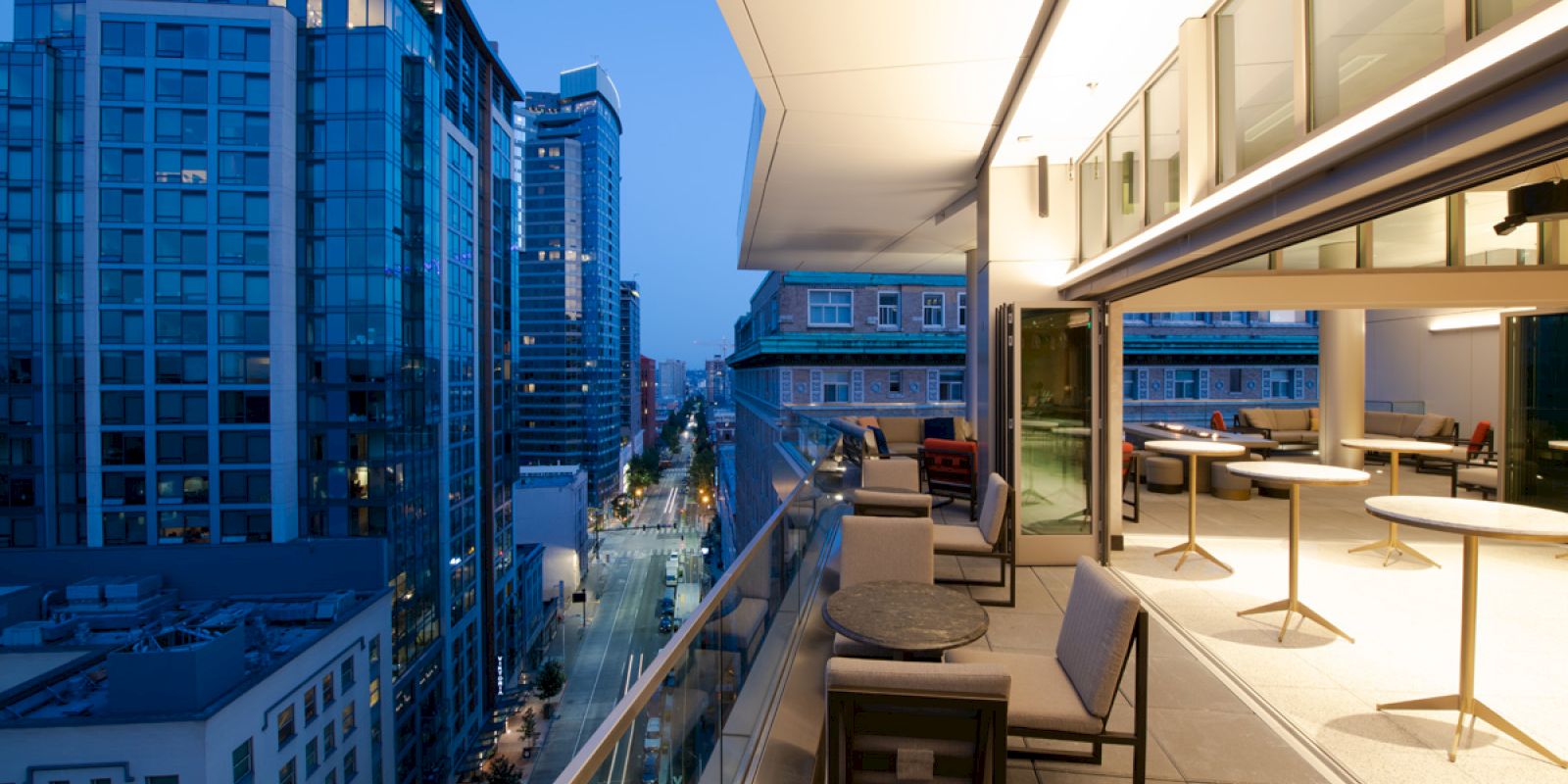 A modern balcony with outdoor seating, overlooking a cityscape with tall buildings at twilight. Some tables and chairs are inside as well.