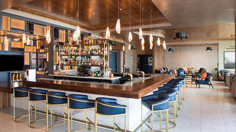 The image shows a stylish bar with a stocked liquor display, blue high stools, pendant lights, and a cozy lounge area in the background.