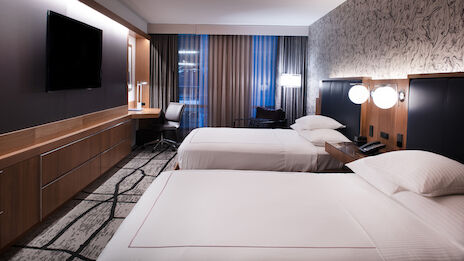 The image shows a modern hotel room with two double beds, a large flat-screen TV, a work desk, and bedside lamps, featuring a contemporary design.