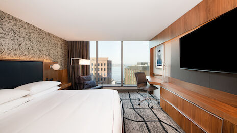 The image shows a modern hotel room with a large bed, a desk, a chair, a television, and a large window offering a city view.