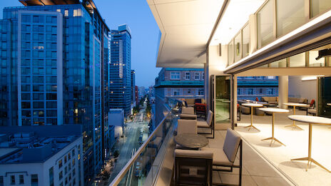 The image shows a modern cityscape at dusk with a tall building and a balcony featuring tables and chairs overlooking the street below.