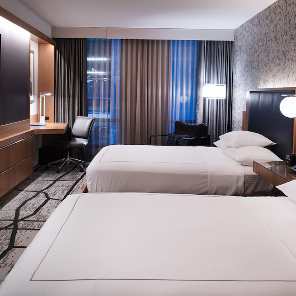 The image shows a modern hotel room with two beds, a TV, a desk with a chair, and ambient lighting. The decor is sleek with a patterned wall.