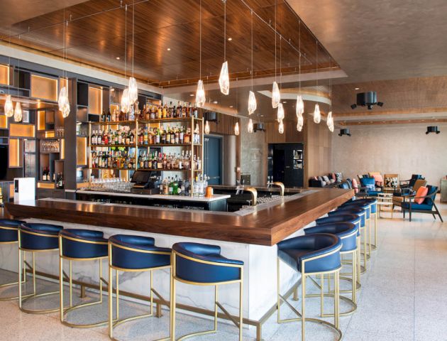 The image shows a modern bar with a wooden counter, blue stools, hanging lights, and a stocked bar shelf, set in a stylish, open space.
