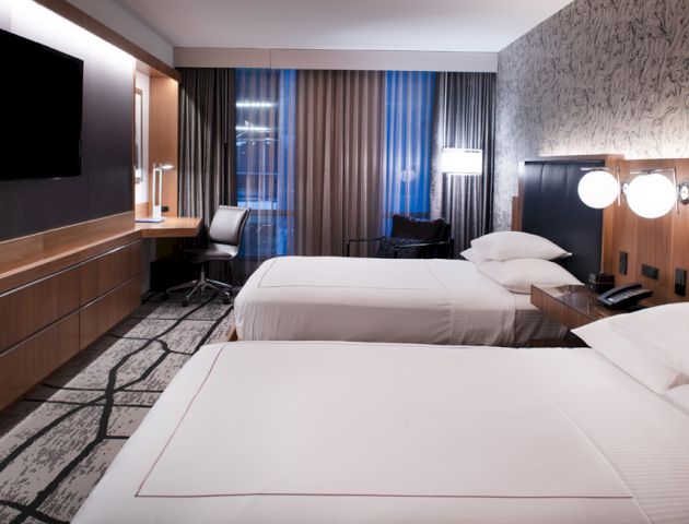 The image shows a modern hotel room with two neatly made beds, a wall-mounted TV, a desk with a chair, and contemporary lighting.