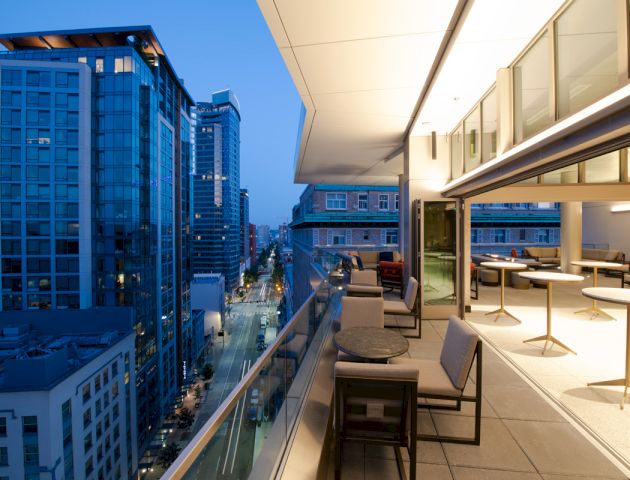 A modern balcony with seating and tables overlooks a cityscape of high-rise buildings during the evening, showcasing a vibrant urban setting.
