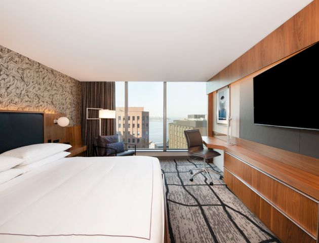 This image shows a modern hotel room with a king-sized bed, a large TV, a desk and chair, and a view of the city through a large window.