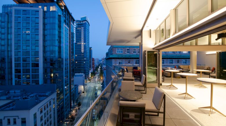 A modern cityscape at dusk, viewed from an open patio with tables, chairs, and a glass balustrade. Tall buildings line the illuminated street.