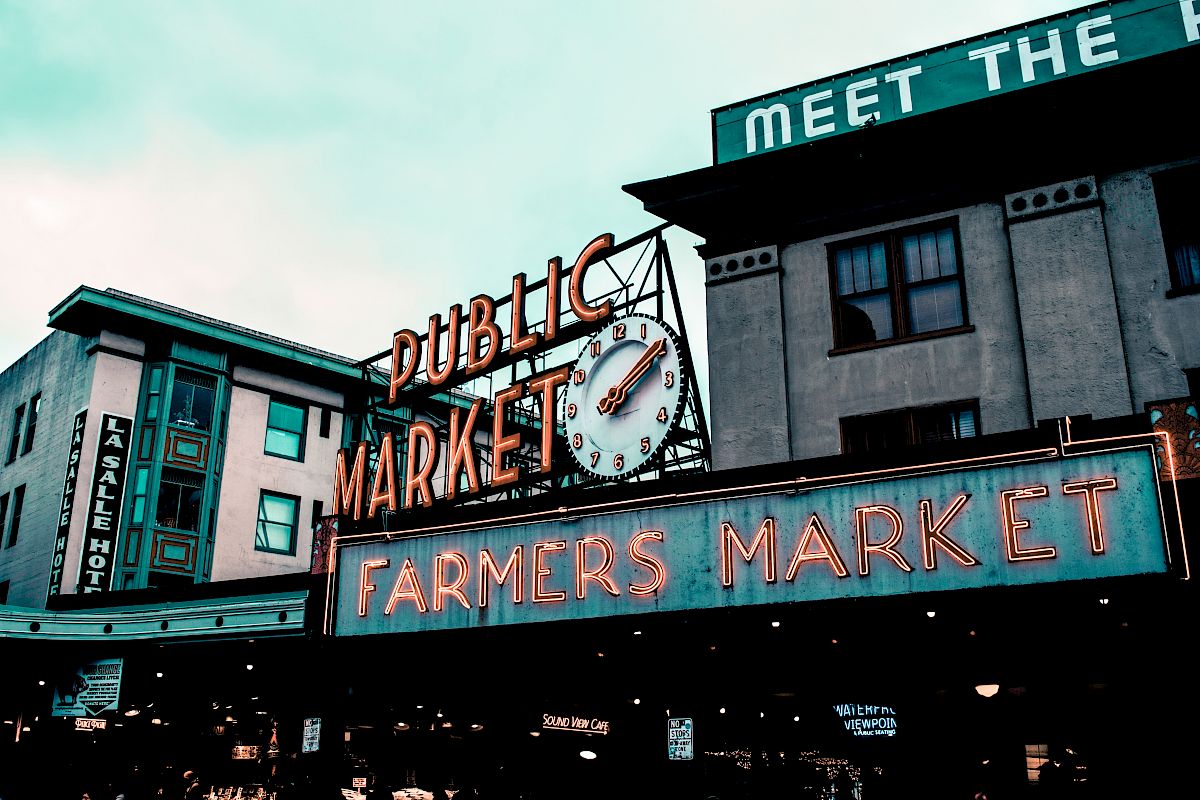 The image shows a vibrant sign reading "Public Market, Farmers Market" with an iconic clock. Buildings and a sign for a hotel are in the background.