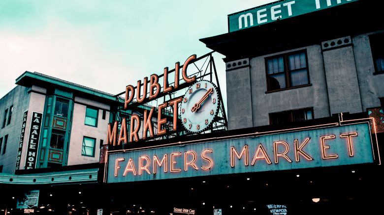 The image shows a vibrant sign reading "Public Market, Farmers Market" with an iconic clock. Buildings and a sign for a hotel are in the background.