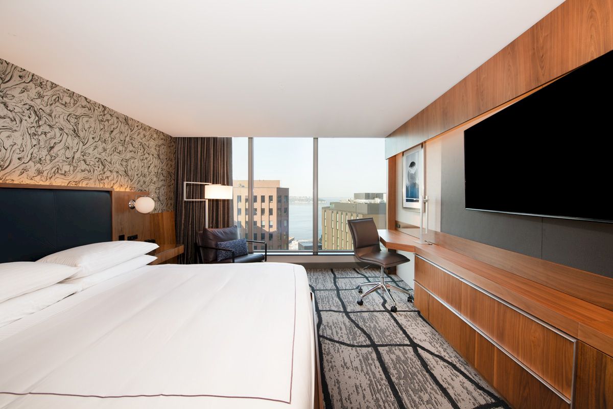 A modern hotel room with a bed, TV, desk, chair, and large window overlooking buildings and water in the distance, decorated with wood and patterns.