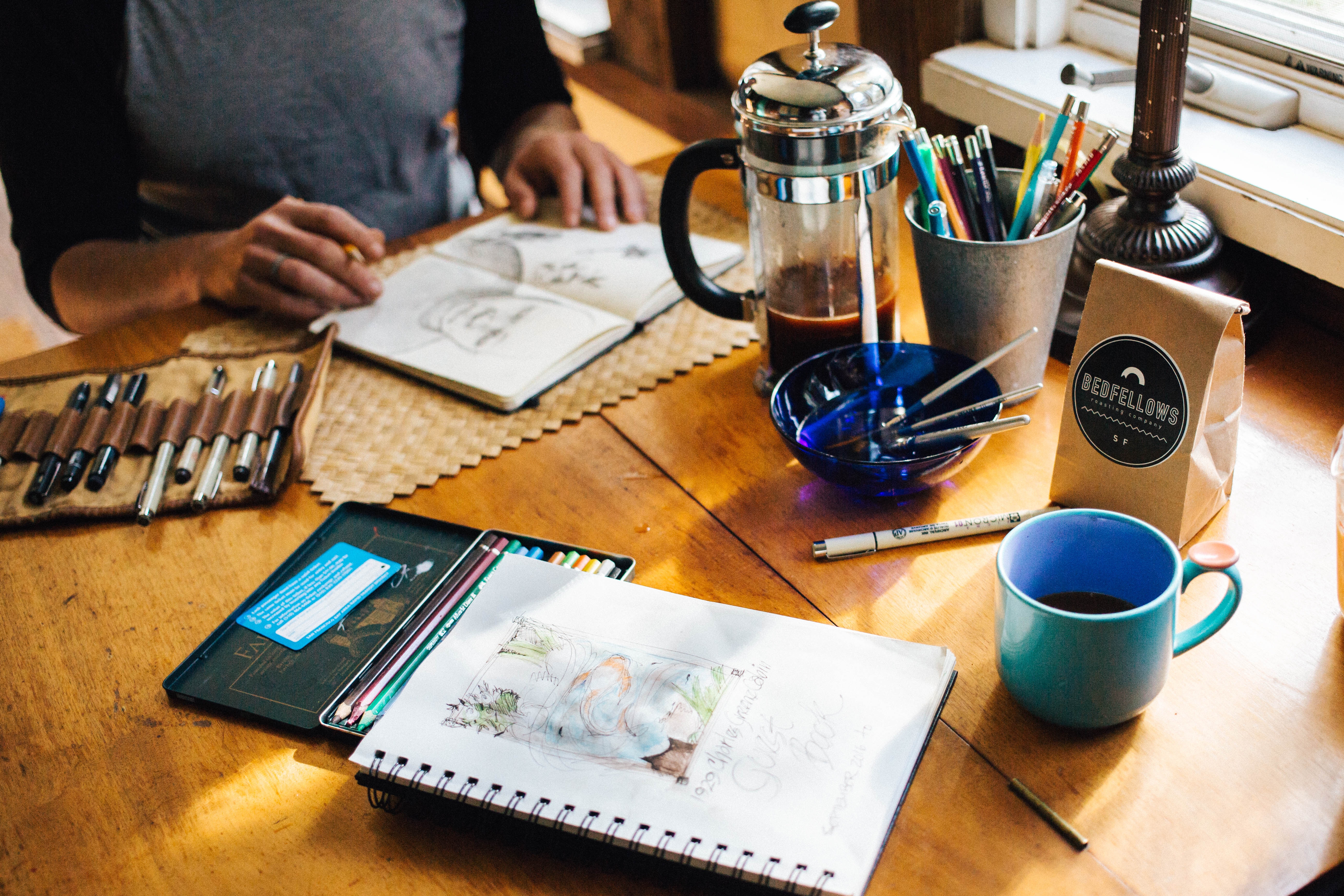 A person sketches at a table with a French press, coffee bag, a blue cup, and various art supplies, including colored pencils, a notebook, and sketches.