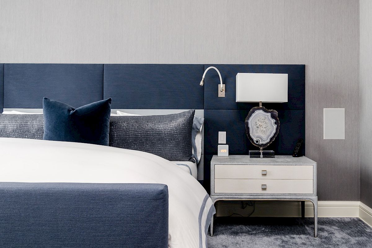 The image shows a modern bedroom with a blue upholstered bed, white and blue bedding, a bedside table with a lamp, and a decorative item.