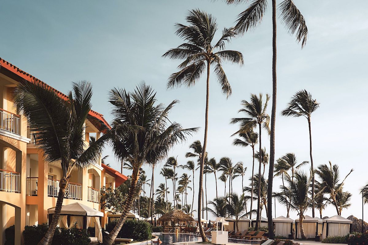 The image shows a luxurious poolside area with lounge chairs and tall palm trees next to a resort building under a clear blue sky.